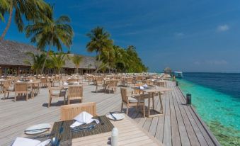 a wooden deck with tables and chairs , palm trees , and the ocean in the background at Vilamendhoo Island Resort & Spa