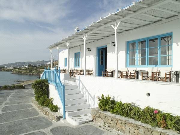 a white building with blue doors and windows , situated near a body of water , with several chairs and umbrellas placed around the area at Mykonos Beach Hotel