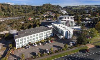a large white building with a green roof is surrounded by trees and other buildings at Rydges Rotorua, an EVT Hotel