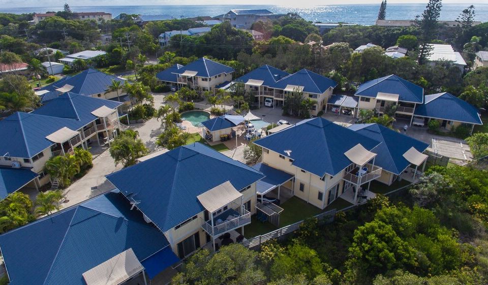 aerial view of a resort with multiple buildings , including blue roofs , surrounded by trees and a beach at Hastings Cove Holiday Apartments