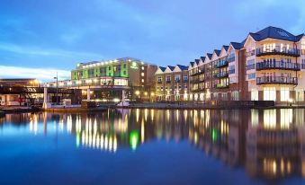 a large building with a green sign is reflected in the water at night , creating a picturesque scene at Holiday Inn London - Brentford Lock