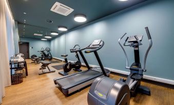 a well - lit gym with various exercise equipment , including treadmills and stationary bikes , on wooden flooring at NH Collection Antwerp Centre