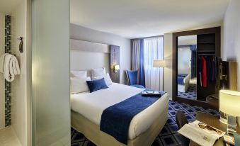a large bed with blue and white linens is in a hotel room with a mirror , nightstand , and window at Mercure Chartres Cathedrale