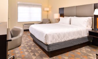 a large bed with white sheets is in a room with a chair , lamp , and window at Holiday Inn & Suites Dallas-Addison
