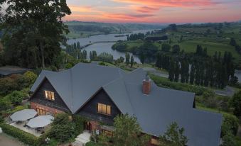 a large house with a wooden exterior is situated in a rural area , surrounded by trees and overlooking a river at Lake Karapiro Lodge