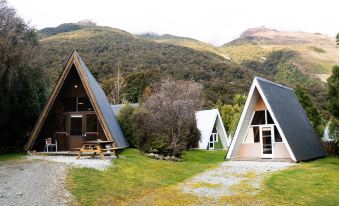 a group of small , triangular wooden houses situated in a grassy area with mountains in the background at Wonderland Makarora Lodge