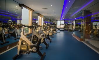 a large , well - lit gym with multiple exercise bikes and treadmills arranged in rows on a blue floor at Ayla Grand Hotel Al Ain