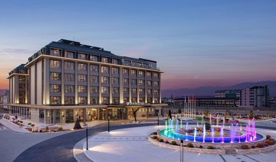 a large hotel with a fountain in front of it , surrounded by snow and trees at DoubleTree by Hilton Skopje