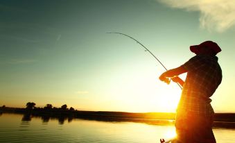 a person is holding a fishing rod over a lake at sunset , casting a fishing line into the water at Camino Real Hotel