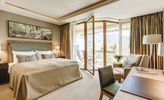a luxurious hotel room with a large bed , a desk , and a view of the mountains outside the window at Althoff Seehotel Uberfahrt