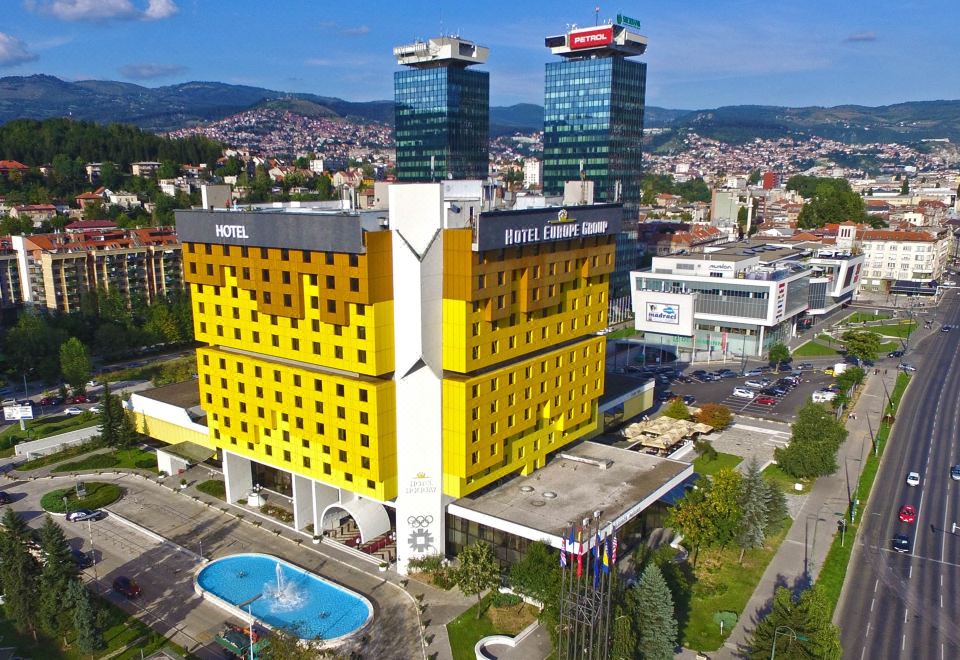 a large yellow hotel building surrounded by a cityscape , with mountains in the background and a swimming pool in the foreground at Hotel Holiday