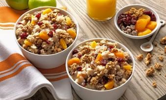 two bowls of granola with fruit and nuts are placed on a wooden table next to an orange juice at Home2 Suites by Hilton Lawrenceville Atlanta Sugarloaf
