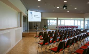 a large conference room with rows of chairs arranged in a semicircle , and a projector screen mounted on the wall at Dialoghotel Eckstein