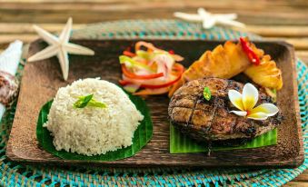 a wooden dining table with a plate of food , including rice , meat , and vegetables , placed on a bamboo tray at Coconut Garden Beach Resort