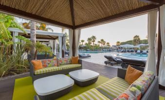 a poolside area with a wooden deck , couches , and tables under a thatched roof , overlooking the pool and palm trees at Conrad Algarve