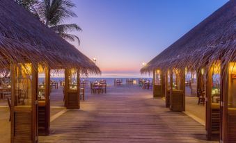 a wooden deck with tables and chairs under thatched roofs , overlooking the ocean at dusk at Meeru Maldives Resort Island