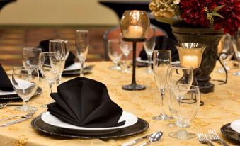 a well - set table with gold tablecloth , black napkins , and wine glasses , along with several other tables and decorations at Hilton Garden Inn Bartlesville
