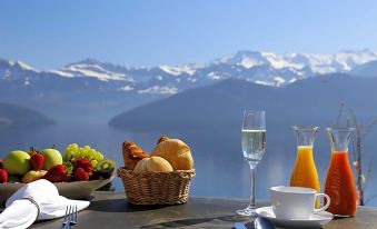a table with a basket of bread , orange juice , and glasses of orange juice is set up in front of a mountainous landscape at Alpenblick Weggis - Panorama & Alpen Chic Hotel
