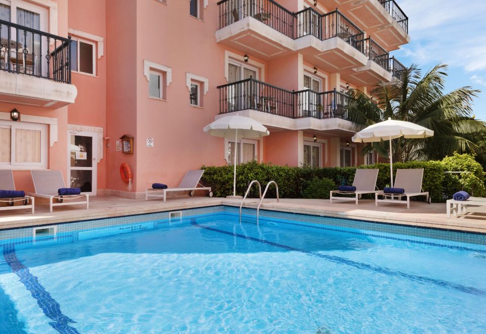 a large swimming pool with white lounge chairs and an umbrella is in front of a pink building at THB Felip - Adults Only
