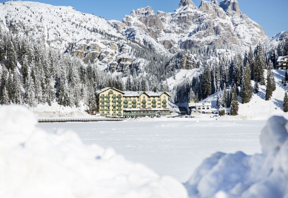 a large building is situated on a frozen lake , surrounded by snow - covered mountains in the background at Grand Hotel Misurina