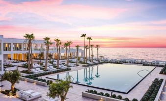 a large outdoor pool is surrounded by lounge chairs and palm trees , with the ocean in the background at Grecotel Lux.ME White Palace
