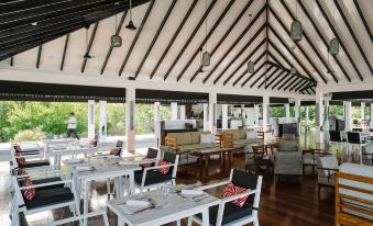 a large , open - air restaurant with white tables and chairs under a wooden roof , surrounded by greenery at Noku Maldives