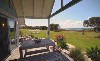 a wooden deck with a dining table and chairs , overlooking a grassy field and the ocean at Bear Gully Coastal Cottages