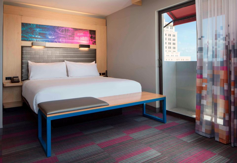 a modern bedroom with a large bed , a blue bench , and a window overlooking the city at Aloft Coral Gables