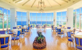 a large , circular dining room with wooden floors and blue chairs , overlooking the ocean through a large window at Kawana Hotel