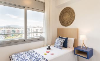 a cozy bedroom with a large window , white walls , and blue accents , featuring a bed with a blue comforter and pillows at Pierre & Vacances Blanes Playa