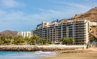 a large , multi - story building is situated on a rocky shoreline near the ocean , with palm trees in the background at Radisson Blu Resort, Gran Canaria