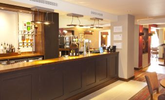 a well - lit hotel bar with a long wooden counter , multiple stools , and a variety of drinks at Premier Inn Rochester