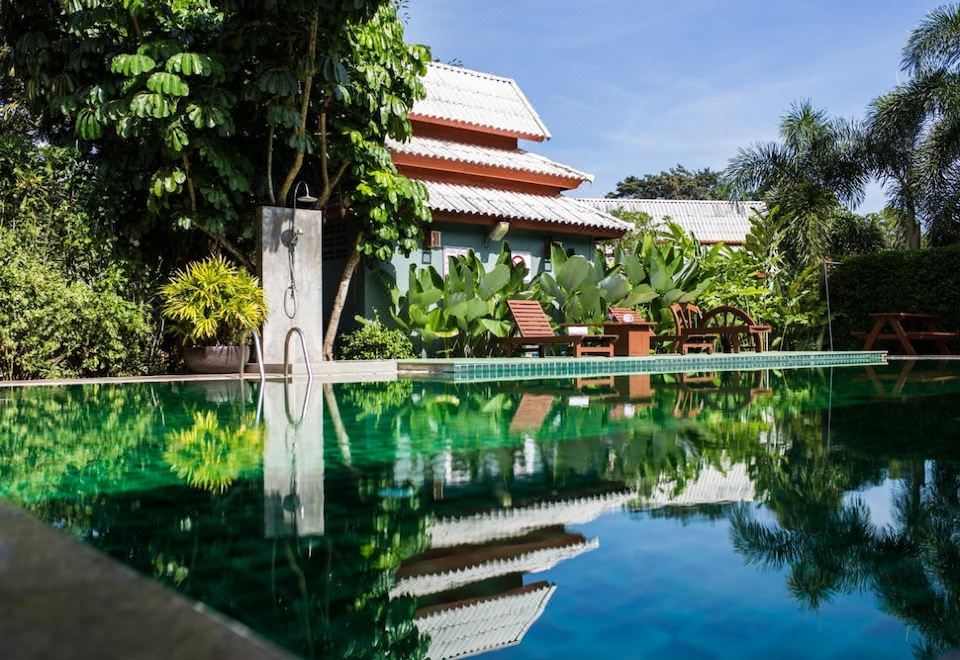 a large swimming pool with a red roof is surrounded by tropical plants and trees , reflecting the surrounding buildings at Poonyamantra Resort