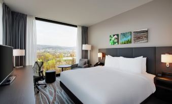 a large , well - made bed is in a room with a chair and a window overlooking a city at Hilton Garden Inn Zurich Limmattal