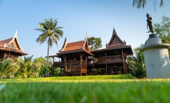a wooden house surrounded by green grass and palm trees , located in a tropical setting at Maikaew Damnoen Resort