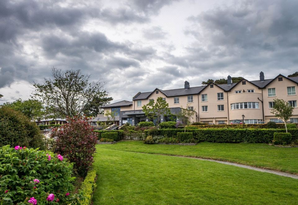 a large building with a red roof is surrounded by greenery and flowers , with a cloudy sky in the background at Arklow Bay Hotel