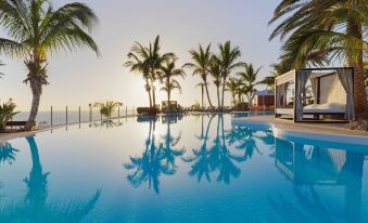 a large outdoor swimming pool surrounded by palm trees , with the sun setting in the background at Roca Nivaria Gran Hotel