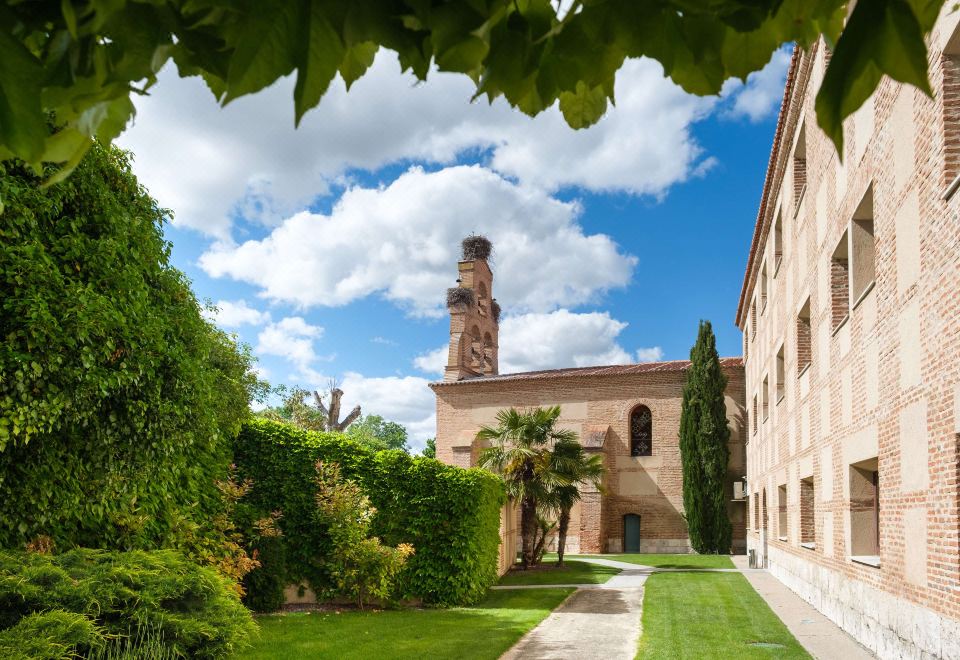 a courtyard with a tall tower in the background , surrounded by green grass and trees at Castilla Termal Olmedo