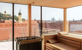a room with a wooden bench and a heater , overlooking a cityscape through the large windows at Hotel Weitzer Graz