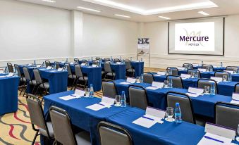 a conference room with rows of blue tables and chairs , each table has a blue tablecloth , water bottles , and a white projector screen at Mercure Belo Horizonte Vila da Serra