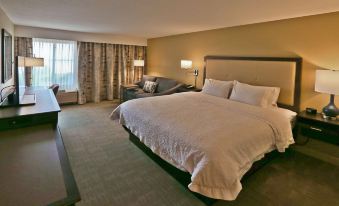 a large bed with a white comforter and pillows is in a room with a couch , lamps , and a window at Hampton Inn Billings