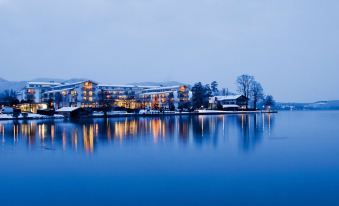 a large building with a clock on its facade is reflected in the water of a lake at Althoff Seehotel Uberfahrt