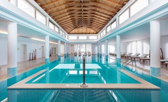 a large indoor swimming pool with a wooden ceiling and white columns , surrounded by lounge chairs and tables at Mitsis Laguna Resort & Spa