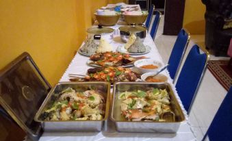 a long table is set with a white tablecloth , two trays of food , and blue chairs in front of it at Sinar Harapan