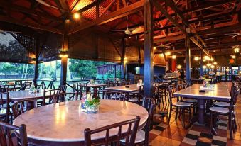 an indoor dining area with wooden tables and chairs , surrounded by a thatched roof and trees at Lavilla by Holiday Villa Cherating
