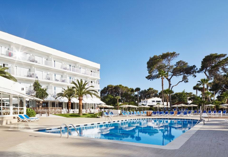 a large white hotel with a swimming pool , umbrellas , and palm trees in front of it at Globales Cala Blanca