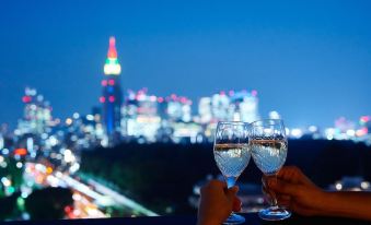 two people are toasting with wine glasses , one of them is holding a clear glass in front of a city skyline at Mitsui Garden Hotel Jingugaien Tokyo Premier
