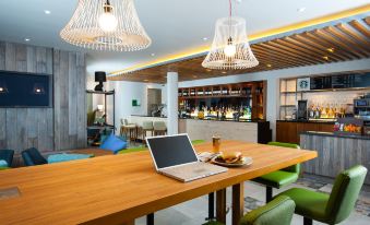 a wooden dining table with a laptop placed on it , surrounded by chairs and a bar in the background at Holiday Inn Salisbury - Stonehenge