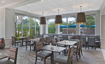 a modern dining area with wooden tables and chairs , surrounded by large windows that offer views of the outdoors at Hilton Garden Inn Jackson/Madison