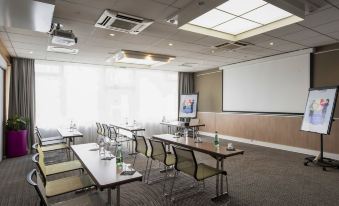 a conference room with rows of chairs and tables , a projector screen , and large windows at Novotel Dijon Sud